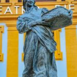Bronze statue of woman with basket in front of colorful building facade in Villaviciosa, Spain.