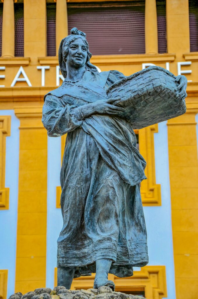 Bronze statue of woman with basket in front of colorful building facade in Villaviciosa, Spain.